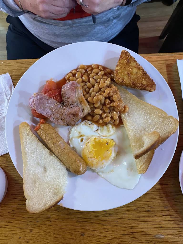 A breakfast plate with egg, bacon, sausage, beans, toast, tomato and hash brown on a wooden table